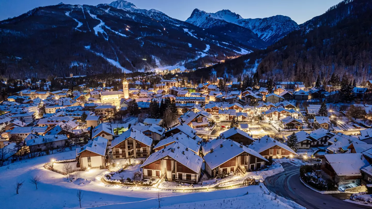 A panoramic view of a village with chalets at night. Snow covered the ground and the roofs. Ski trails on mountains are visible in the distance.