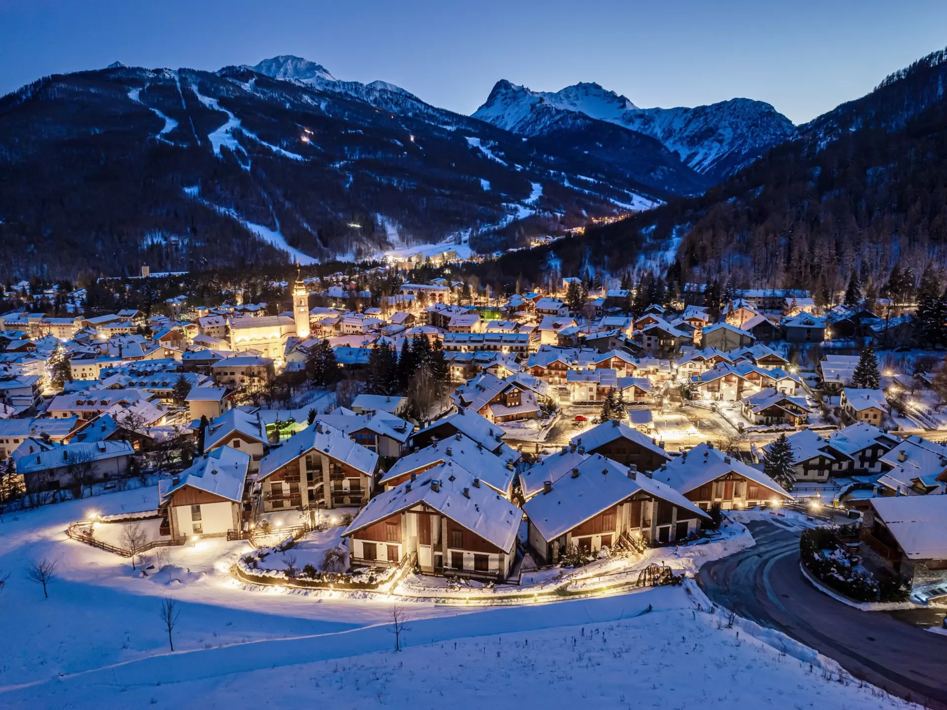 A panoramic view of a village with chalets at night. Snow covered the ground and the roofs. Ski trails on mountains are visible in the distance.