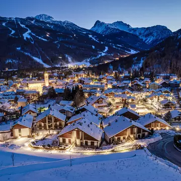 A panoramic view of a village with chalets at night. Snow covered the ground and the roofs. Ski trails on mountains are visible in the distance.
