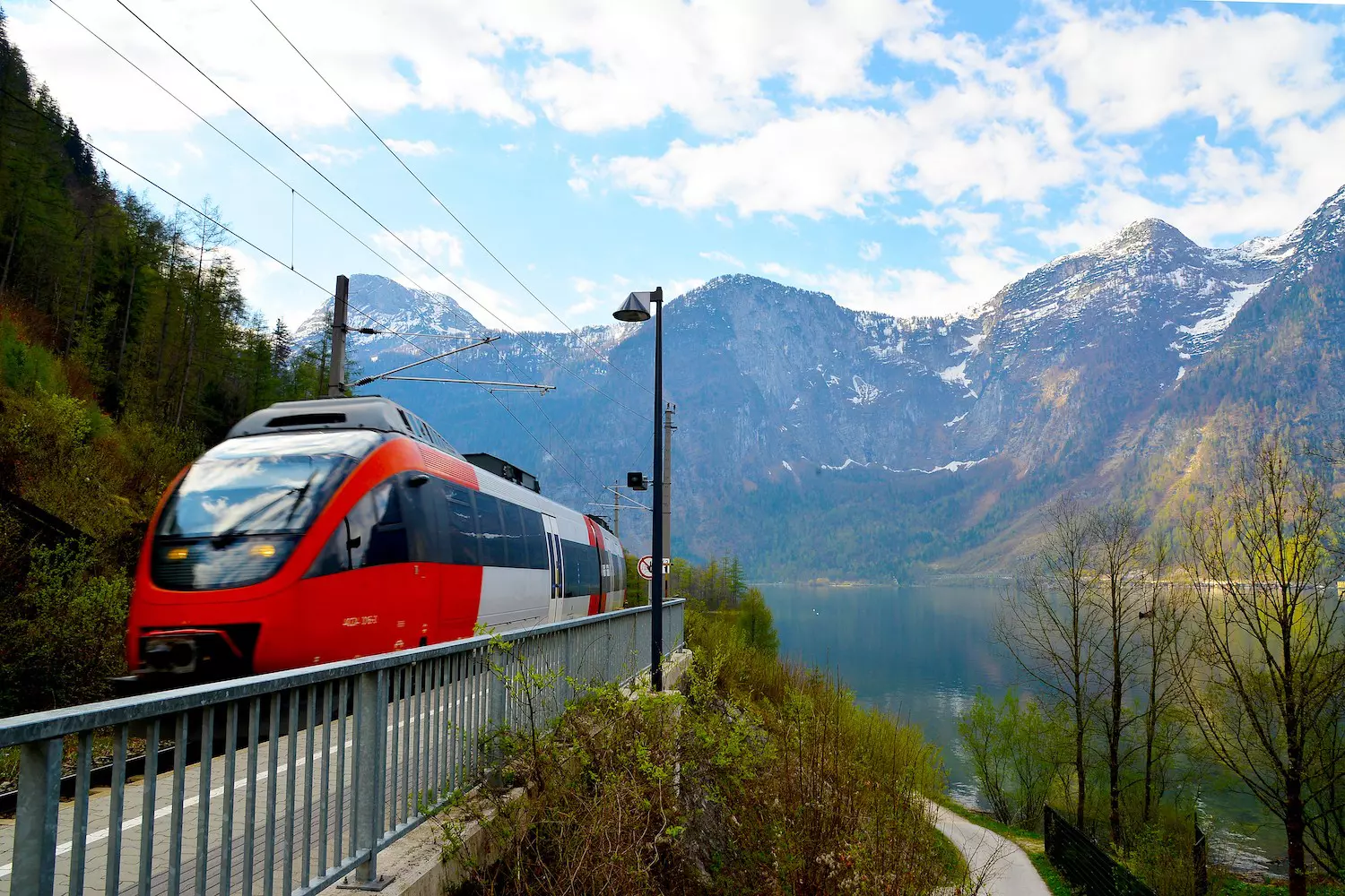 A high-speed train passing the Hallstatt station, on a bridge over water with mountains in the distance