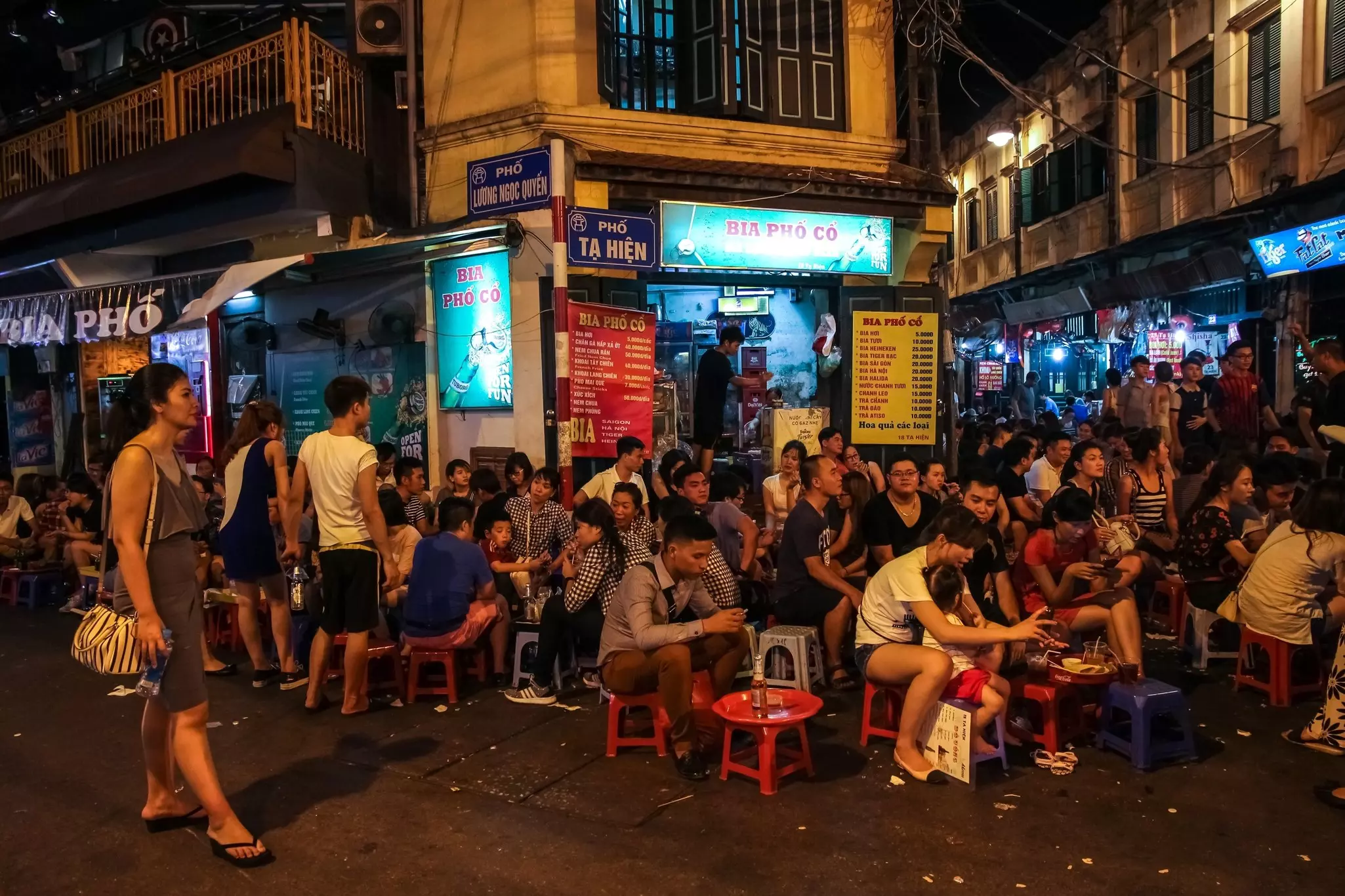 A crowded street corner in the old quarter of a city with people sitting on stools while enjoying drinks at a cafe.