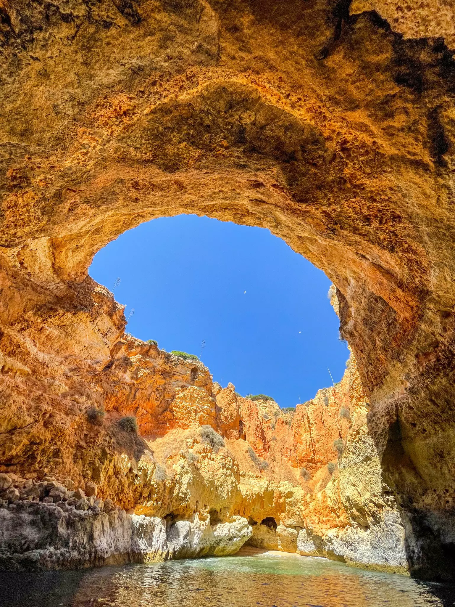 A sea cave with colorful yellow and orange walls. The collapsed cave roof allows a sightline directly to the sky.