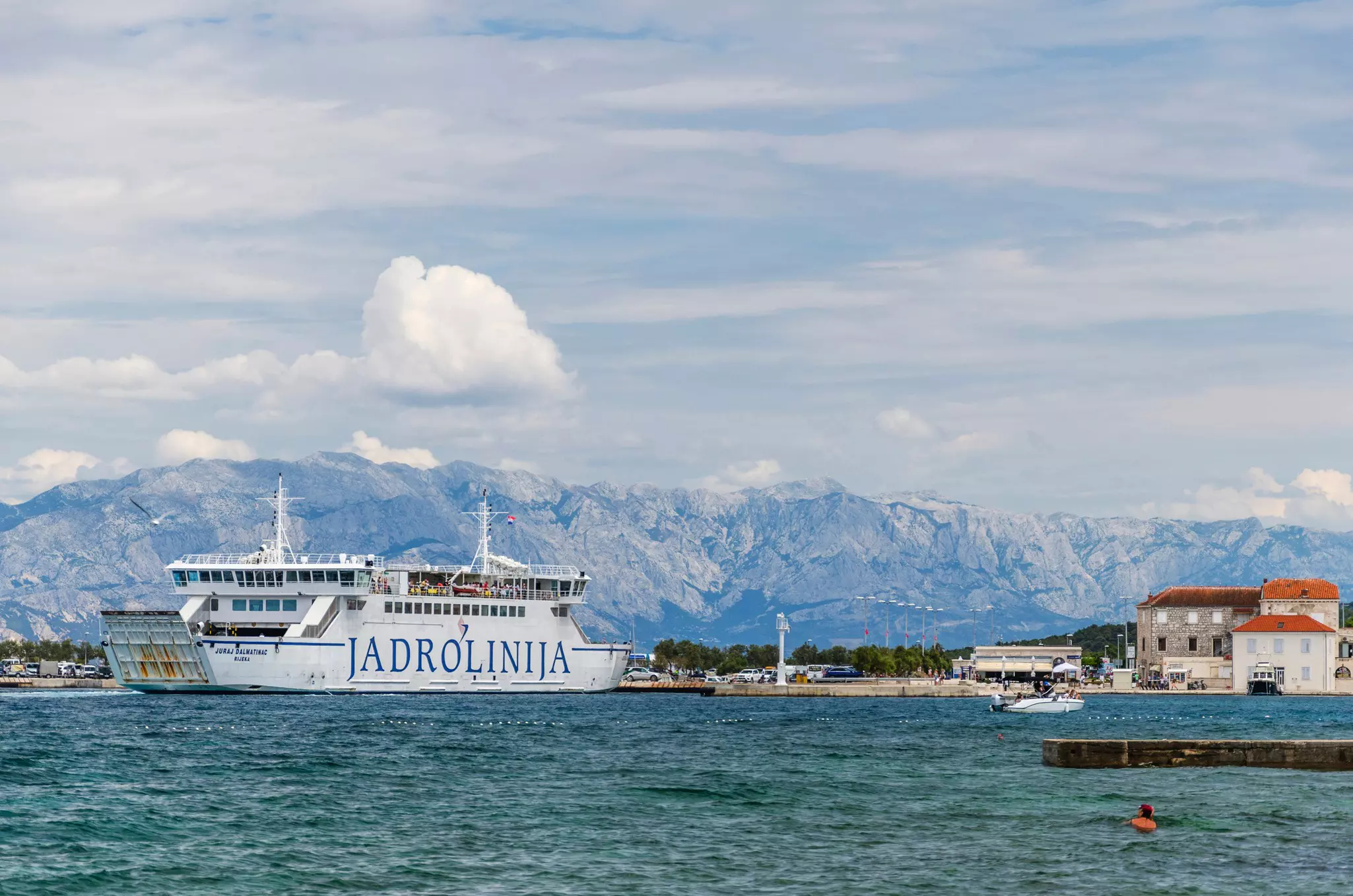 A Jadrolinija ferry departs a port. A ridge of rocky mountains is seen in the distance.