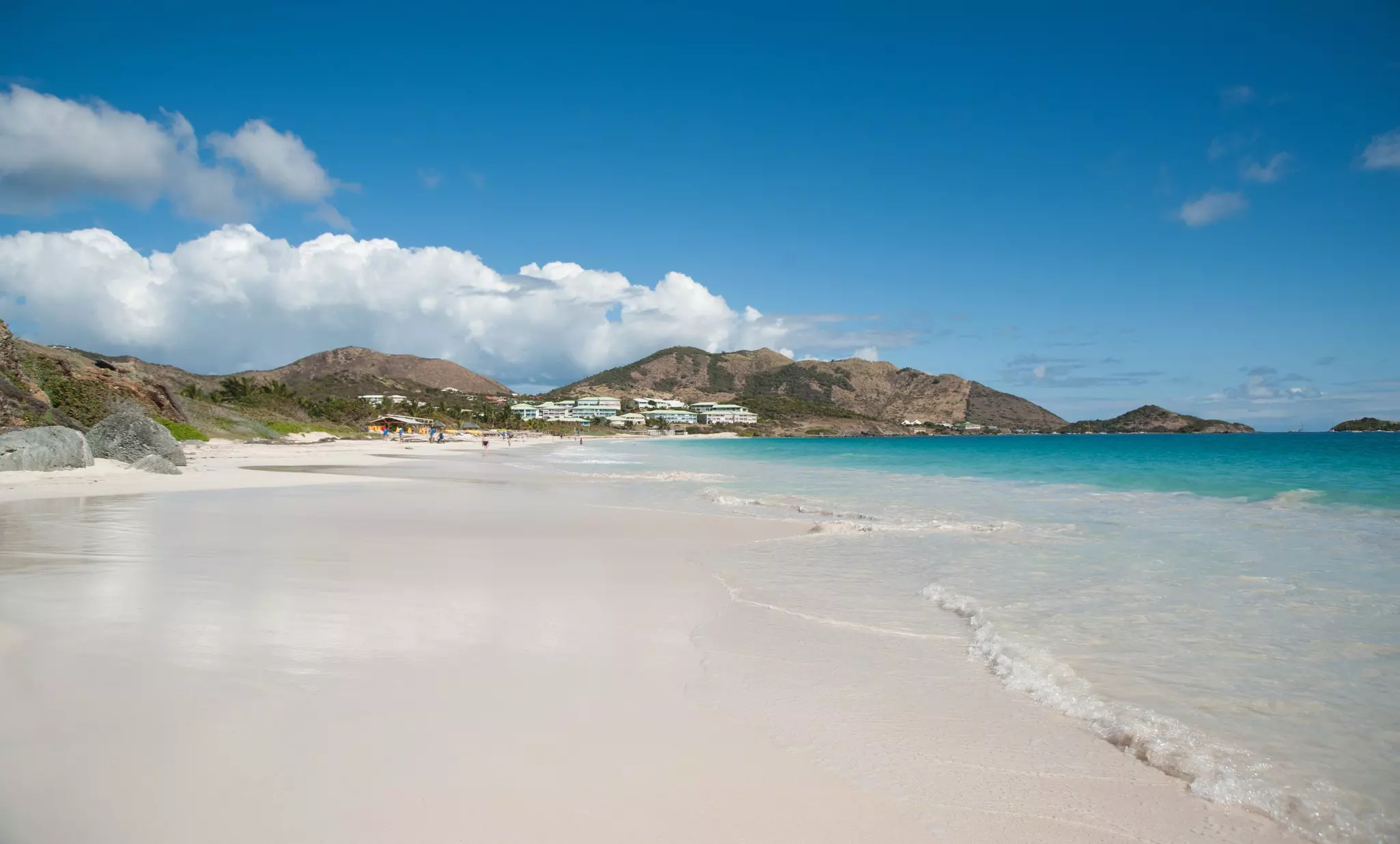 A wide, white sand beach with turquoise water to the right and low hills and buildings in the distance on a sunny day. A few people appear to be on the beach in the very far distance.