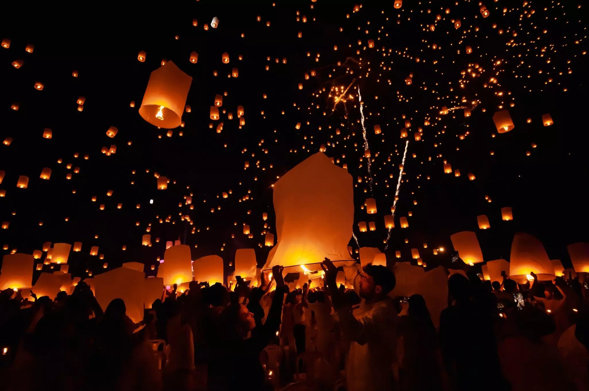 People in silhouette release glowing orange lanterns into the dark night sky.