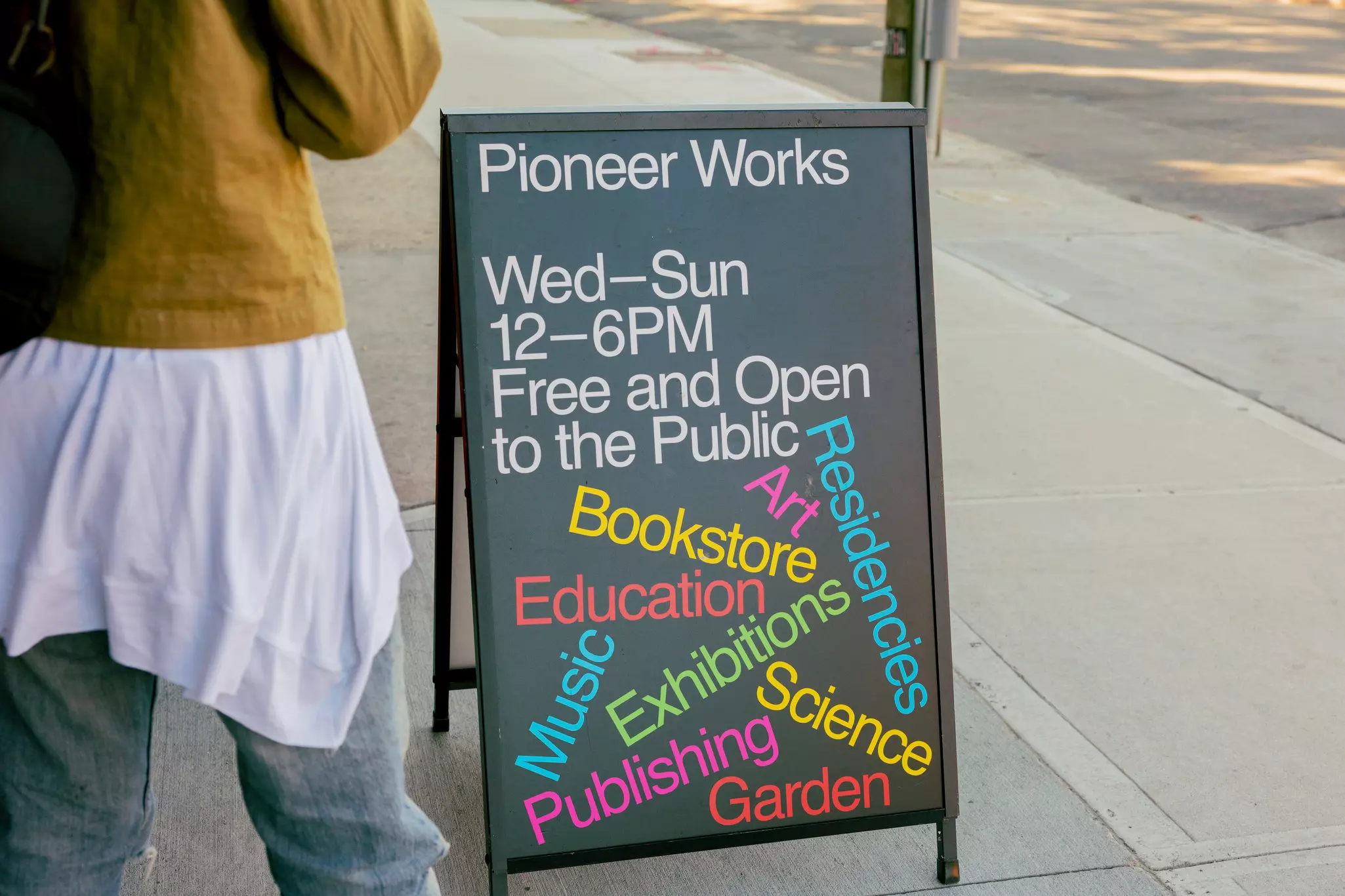 A sidewalk sign for Pioneer Works, with the hours and colorful examples of events.
