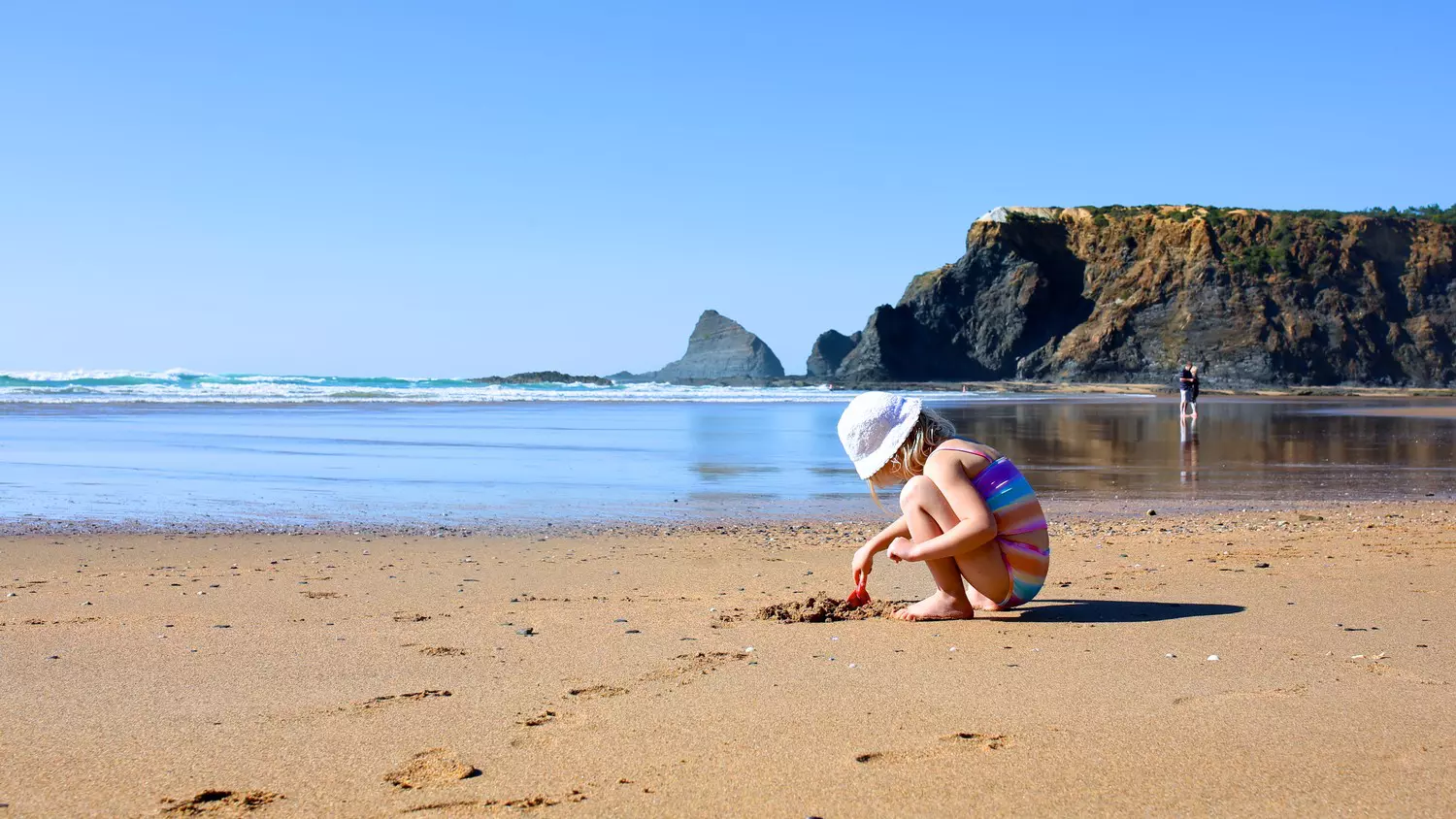 A child playing in the sand at Praia de Odeceixe in the south of Portugal, with cliffs behind.