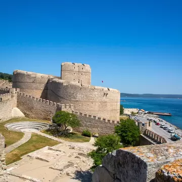 Stone walls and rounded keep of Kilitbahir Castle, with a small amphitheatre and ruins below and a shore road by the strait