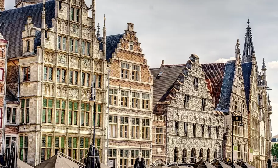People board boats on a waterway in front of Gothic guildhalls with stepped gables.