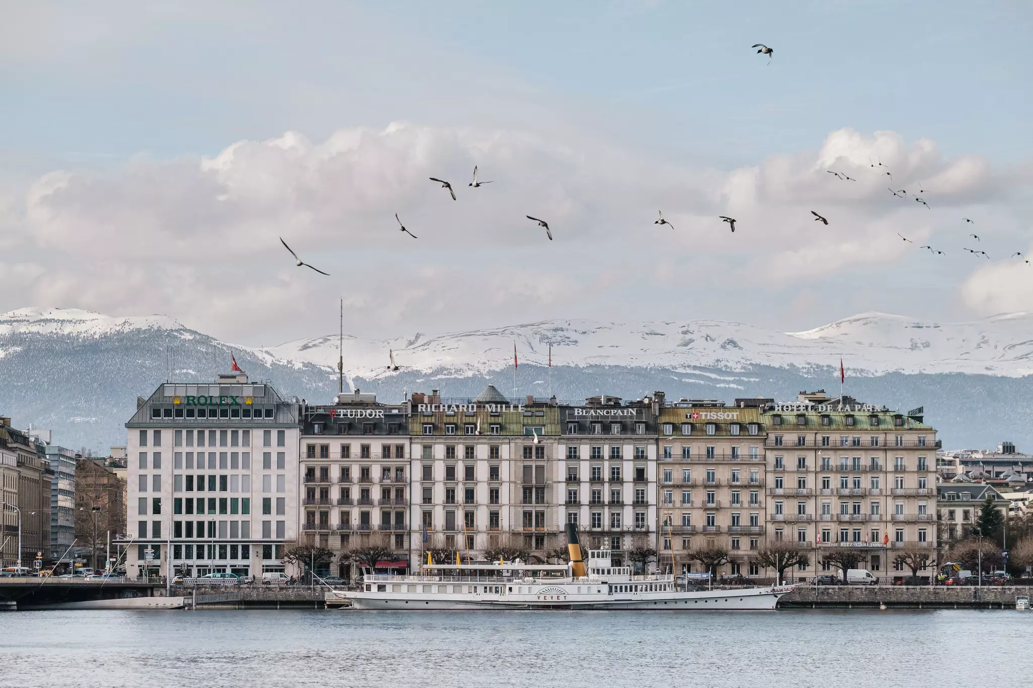 Luxurious buildings line a waterfront passage in a city. Birds fly overhead, a historic steamer is docked along the quay, and snow-capped mountains can be seen rising in the distance.