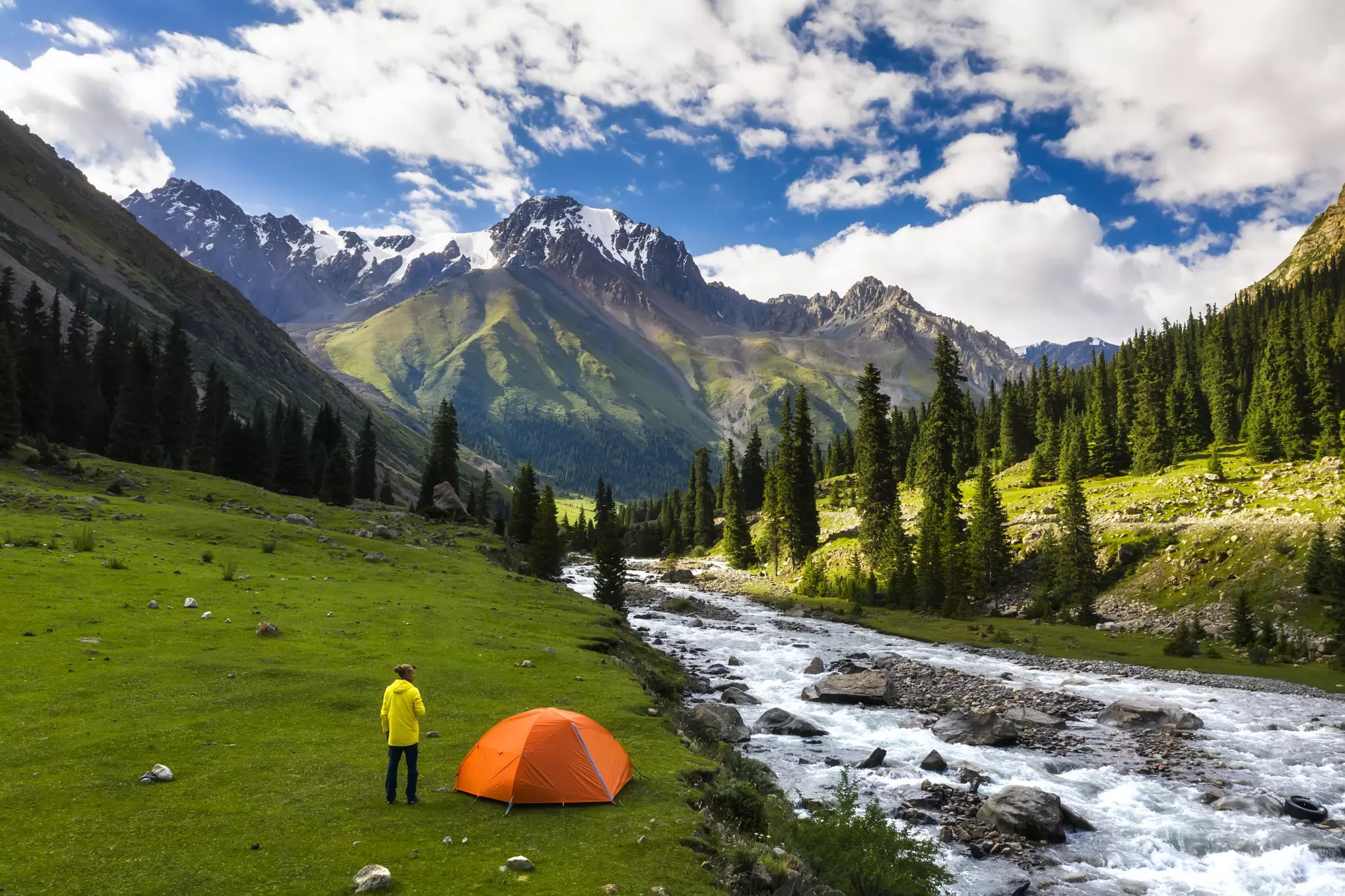 A man stands next to his orange tent in a green mountain valley next to a roaring river. Snow-capped peaks and pine trees are visible in the distance.