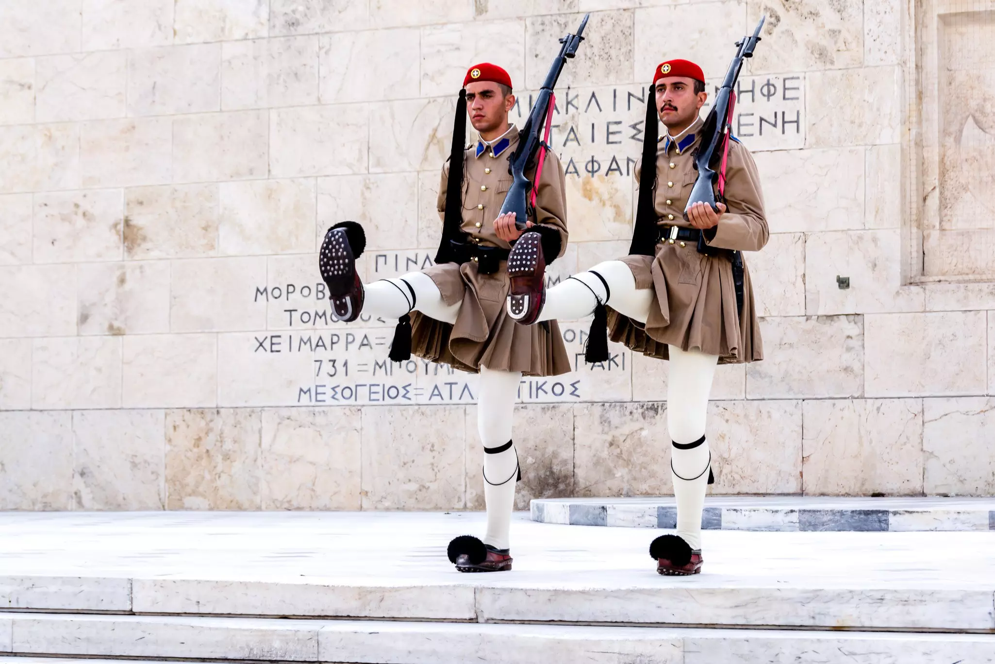 Two guards in traditional costumes kick their legs in formation on the steps of a public building.