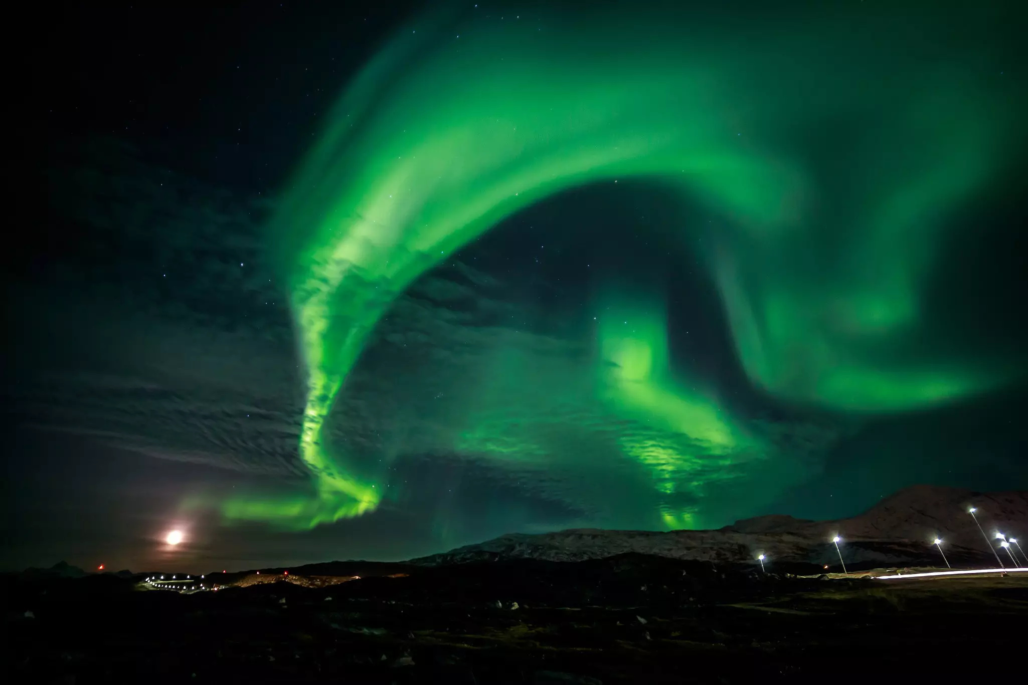 Green northern lights streak the night sky over a dark silhouette of land in Greenland.