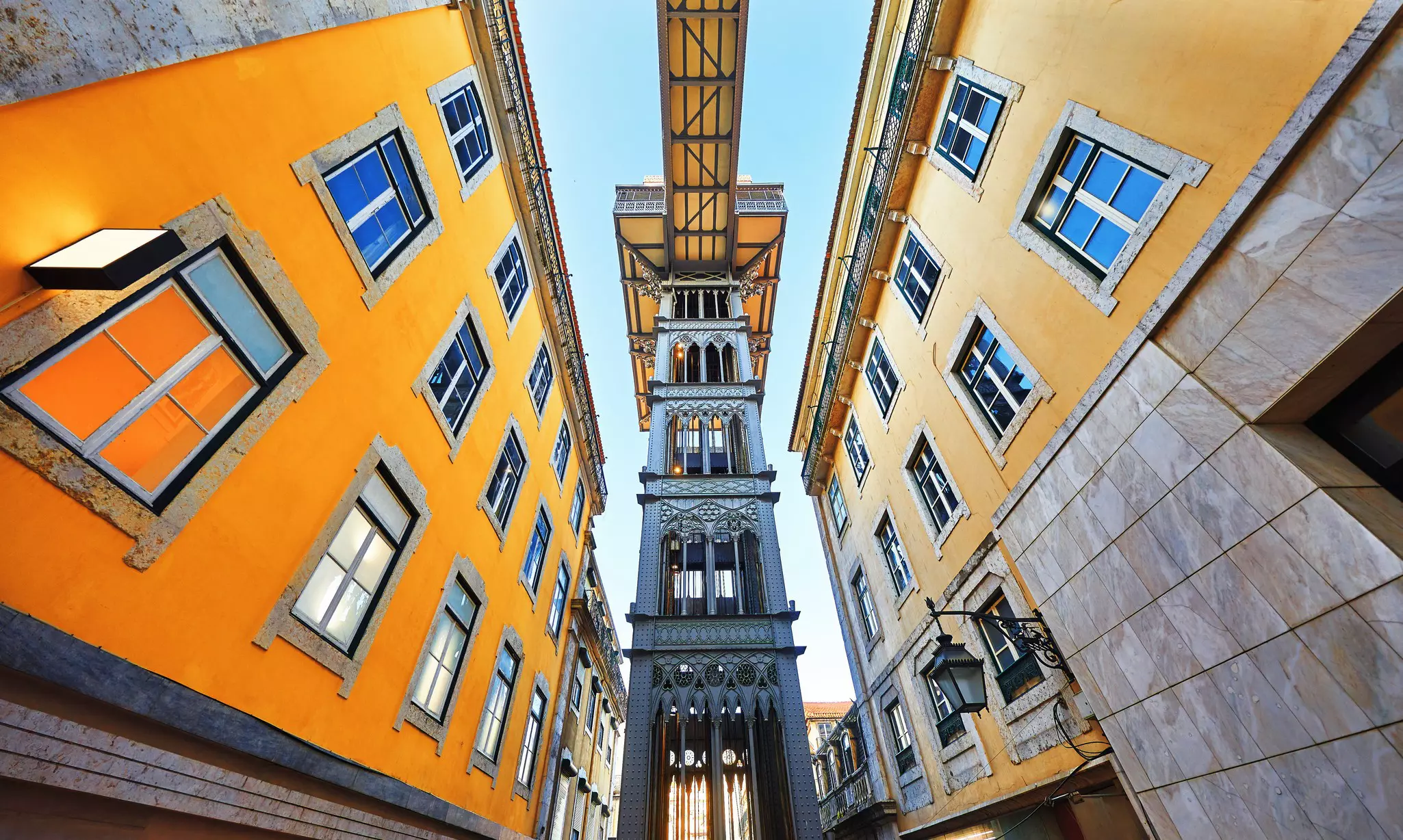 Looking up at the Santa Justa elevator between two yellow buildings on a sunny day.