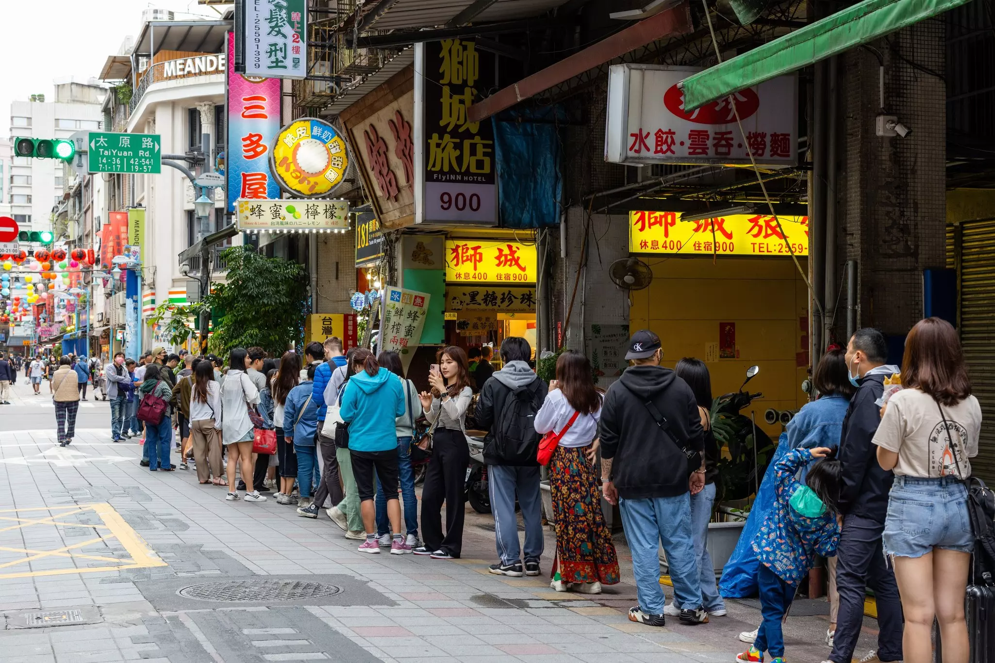 People form an orderly queue in a street as they wait to enter a restaurant. Colorful signs hang from buildings that line the street.