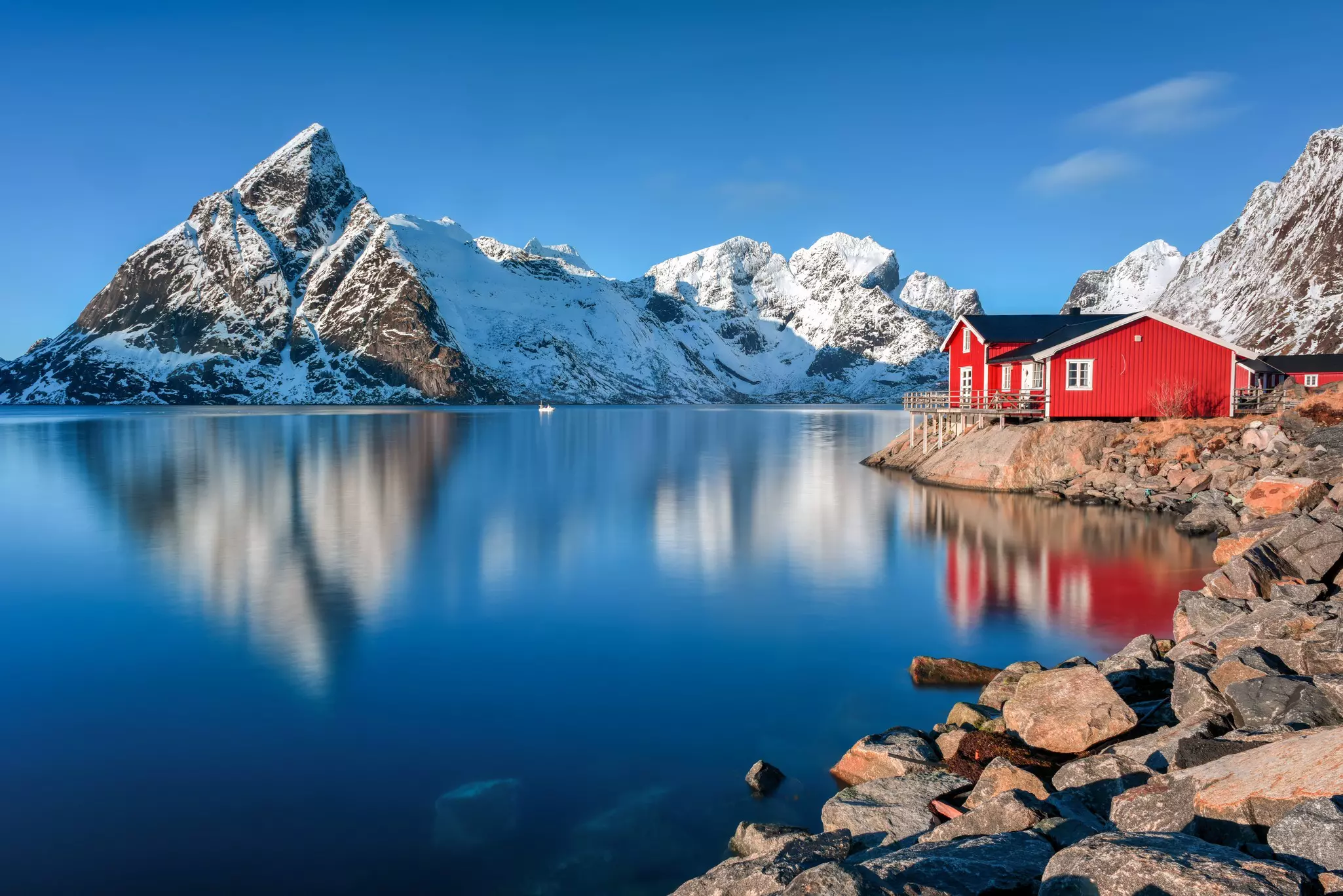 A red wooden building at the edge of a the water with snow-covered peaks rising in the distance.