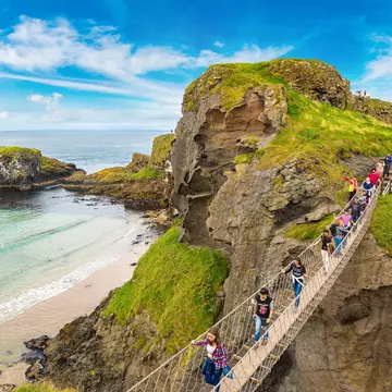 An overhead view of people walking across a narrow bridge that spans a gorge above a beach by a rocky headland.