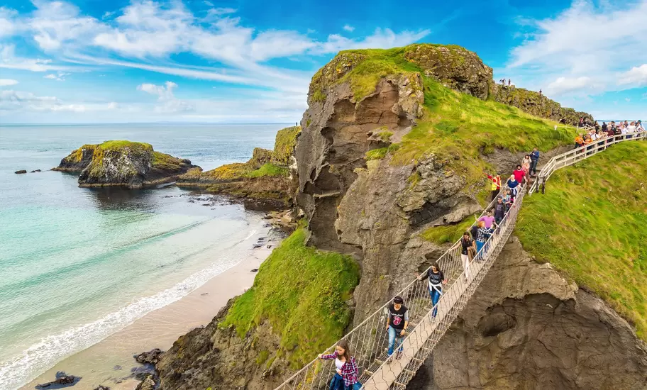 An overhead view of people walking across a narrow bridge that spans a gorge above a beach by a rocky headland.