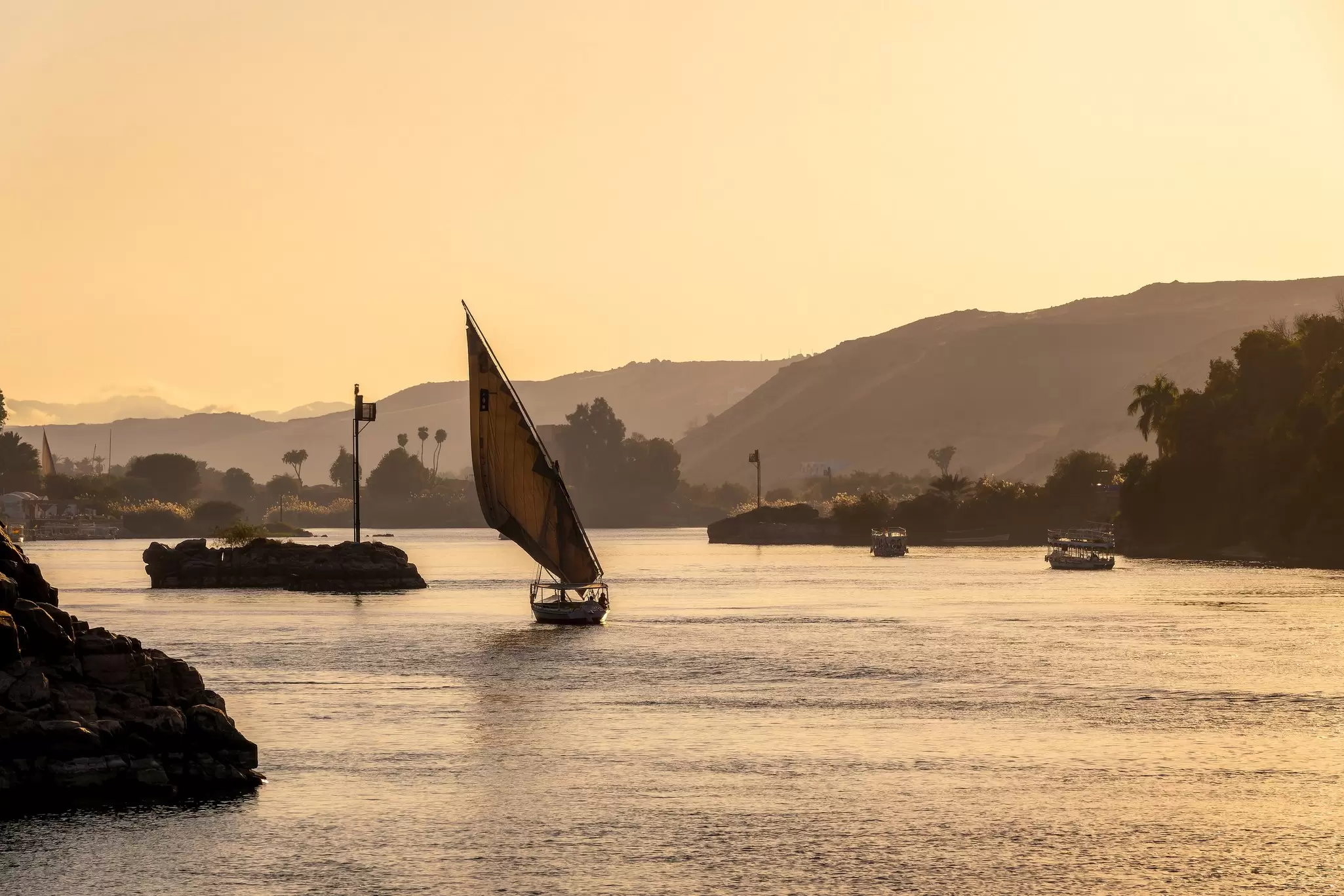 A small sailboat is silhouetted in the light of sunset as it plies a river with hilly banks.