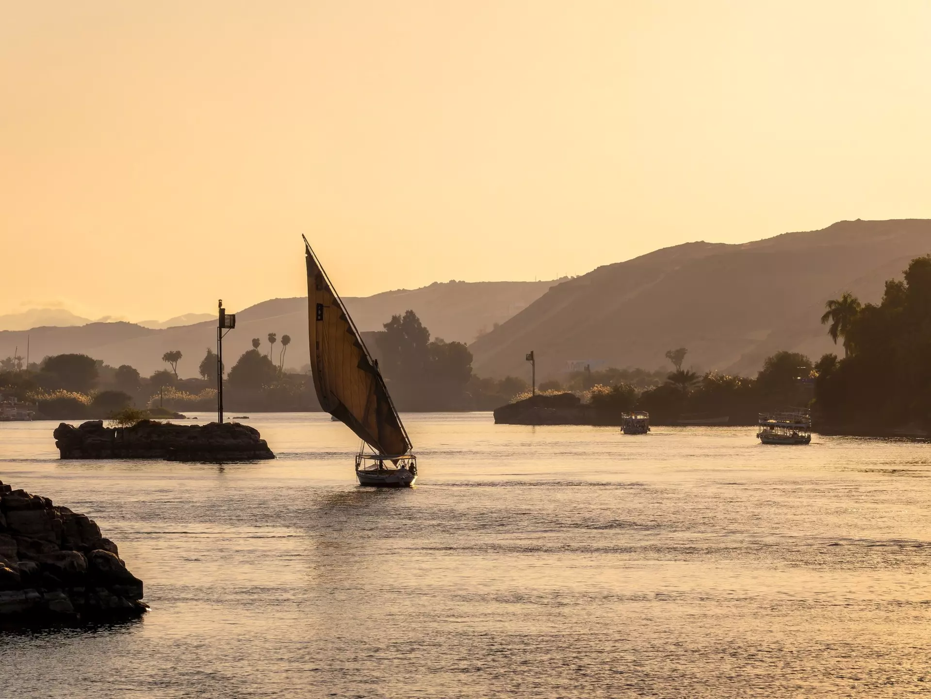 A small sailboat is silhouetted in the light of sunset as it plies a river with hilly banks.