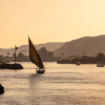 A small sailboat is silhouetted in the light of sunset as it plies a river with hilly banks.