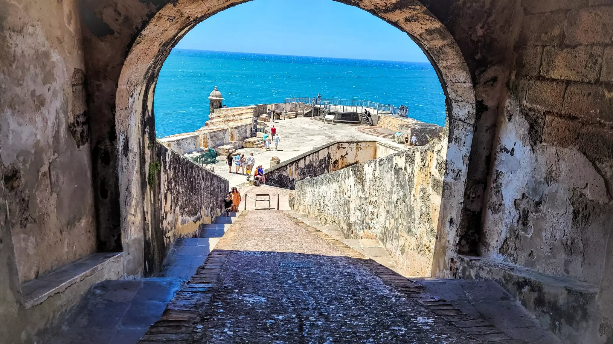 A cobblestone ramp leads down to an open area ringed by the walls of a historic fort by the sea.