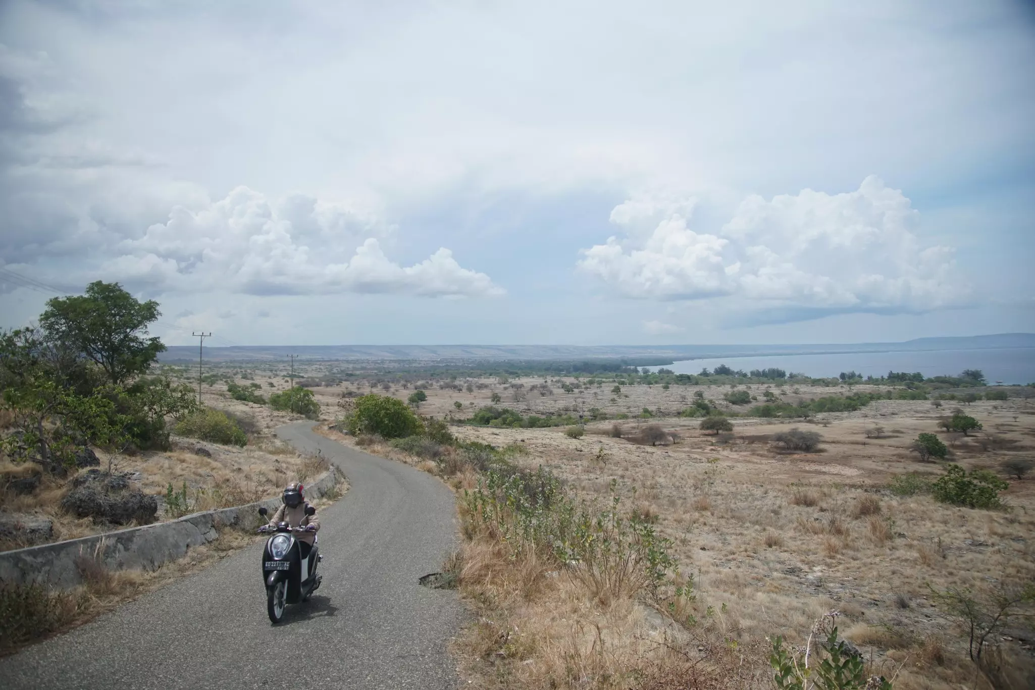A man on a motorcycle drives on a road through a dry, remote landscape. The sea is visible in the distance.