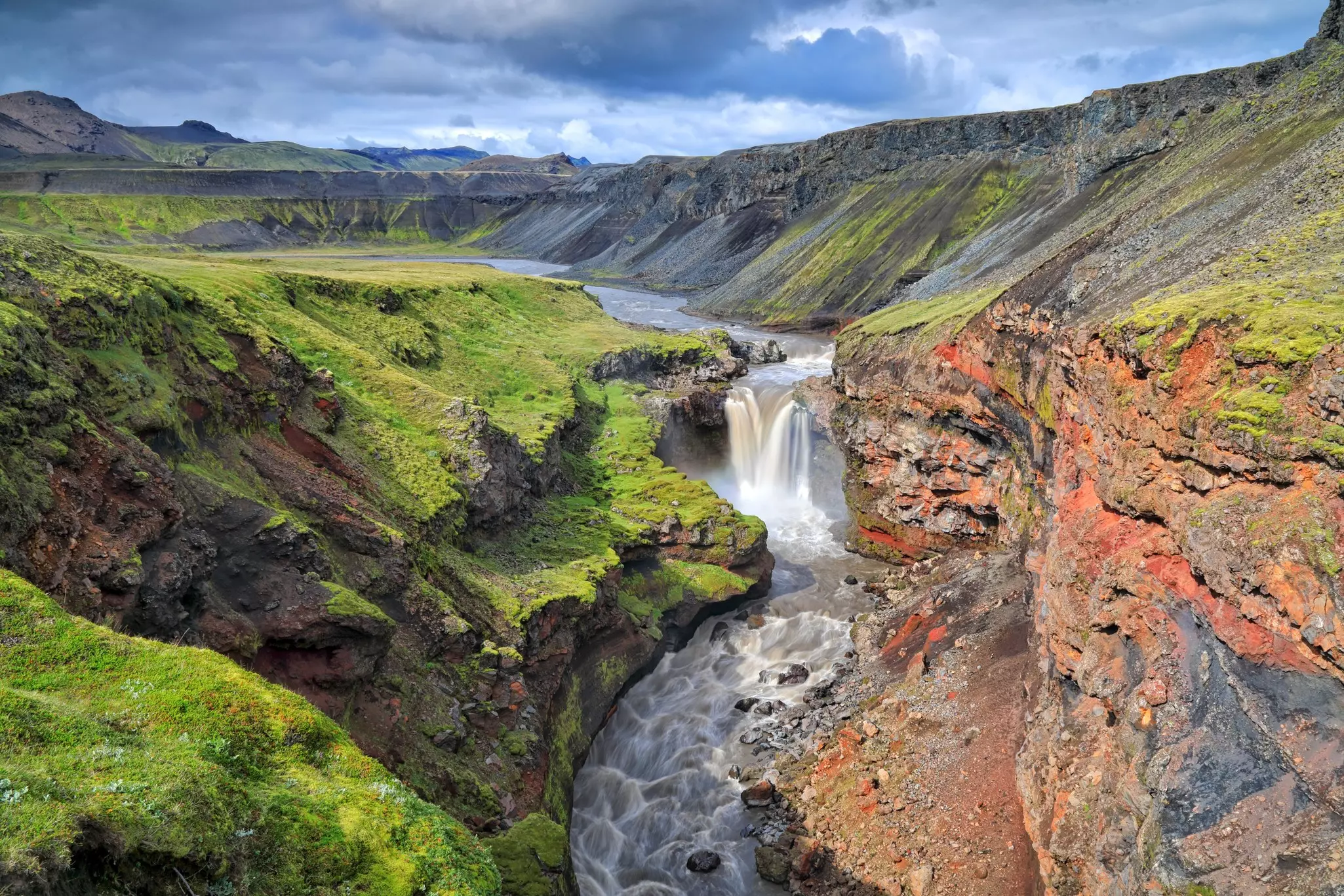 An aerial view of a waterfall in a canyon with multicolored rocks and green vegetation growing on its sides.