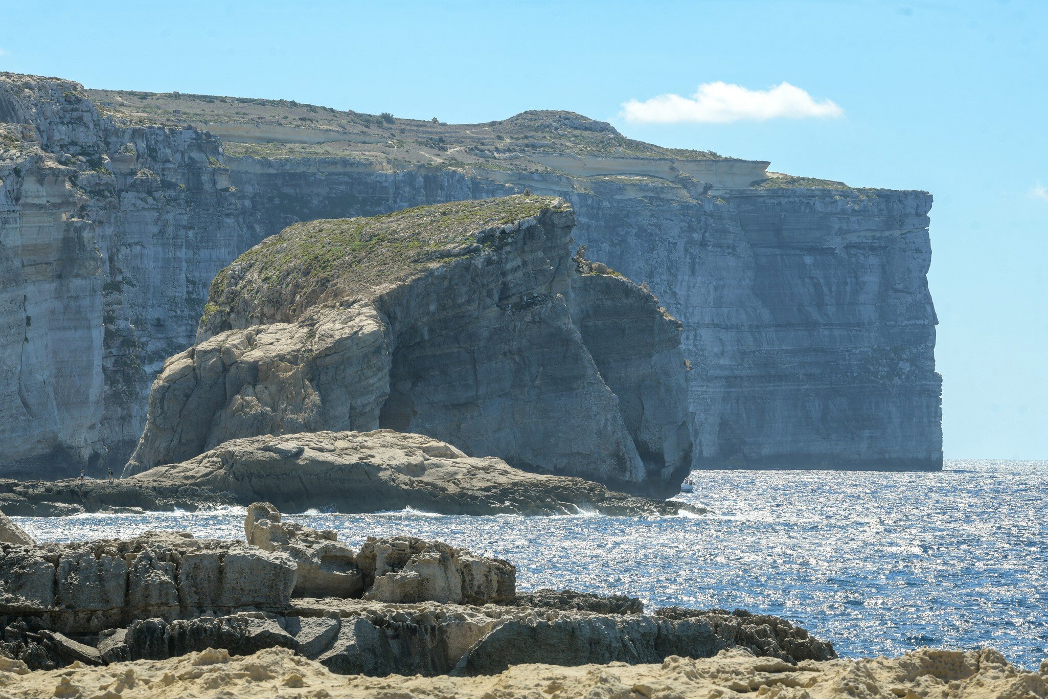 Huge flat-topped rock stack beside towering layered sea cliffs, glittering blue water and eroded rocks in the foreground