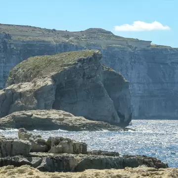 Huge flat-topped rock stack beside towering layered sea cliffs, glittering blue water and eroded rocks in the foreground