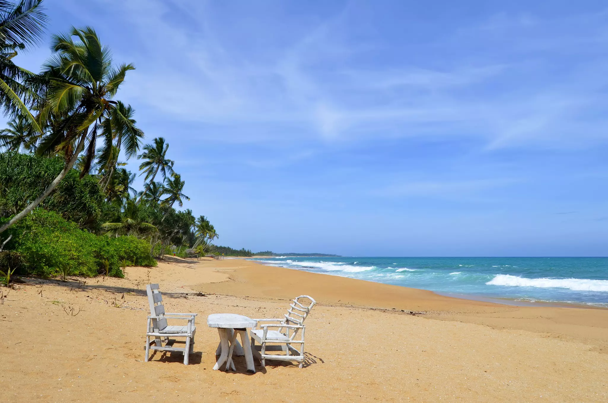 A table and two chairs are set on an empty tropical beach. White-capped waves lap the shore, and palm trees lean into the wind on the shore.