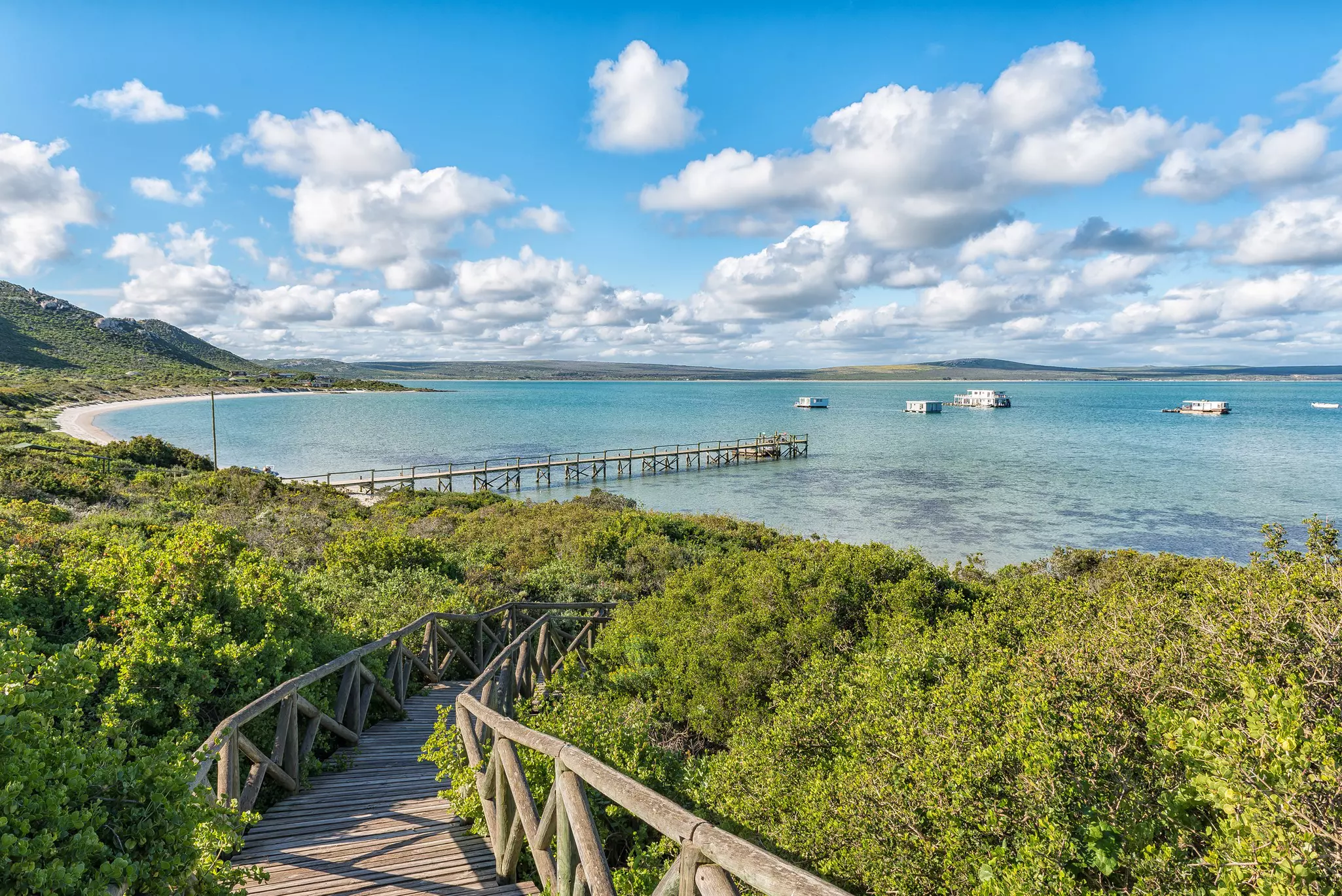 Turquoise waters lapping against a white-sand beach reached by a long wooden boardwalk through bush.