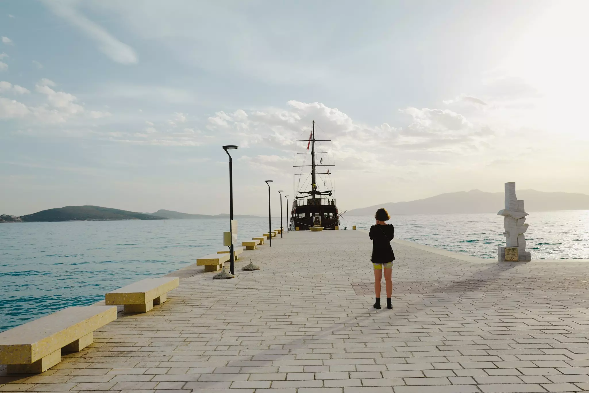 Woman standing alone at a dock in Albania with a boat moored at the end and blue sea around 