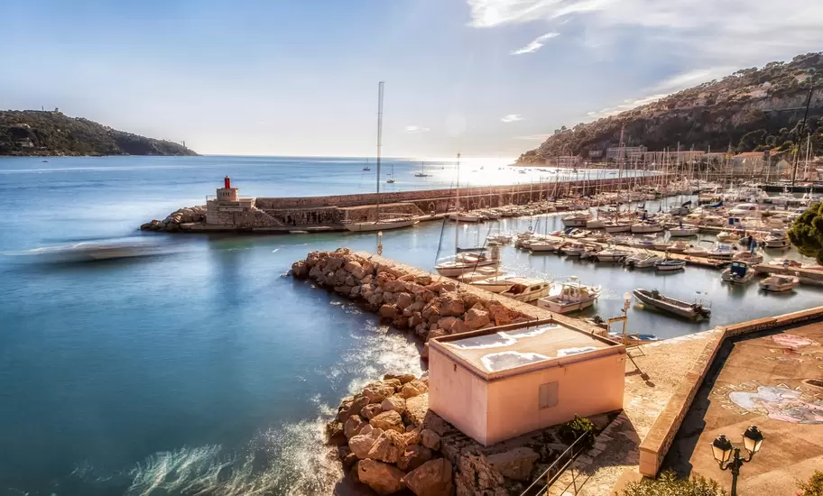 Marina with moored boats and a stone jetty topped by a red lighthouse beside wooded hills on the Côte d'Azur coast