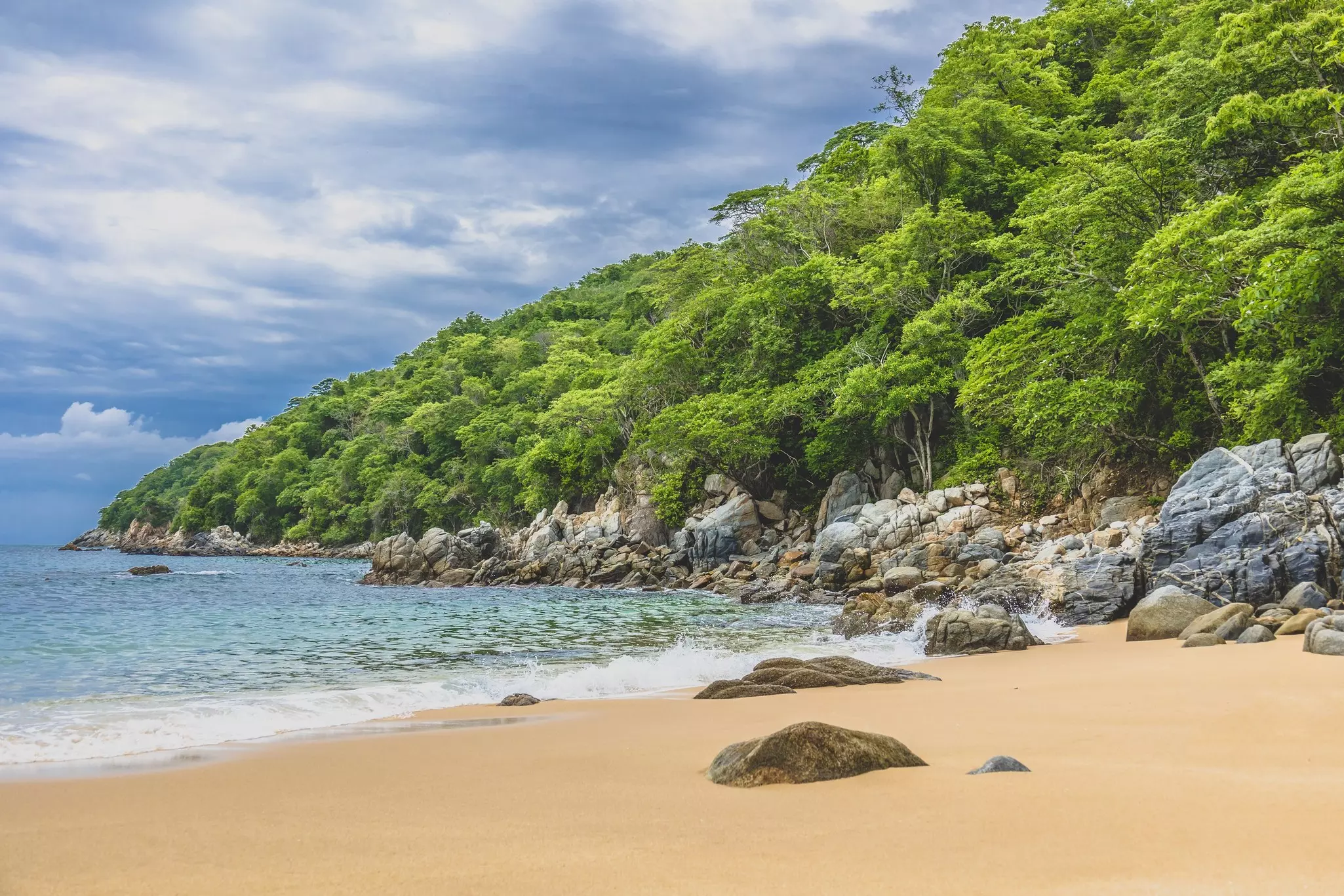 A deserted beach lined by boulders on a cloudy day. Greenery covers the hill rising behind the beach.