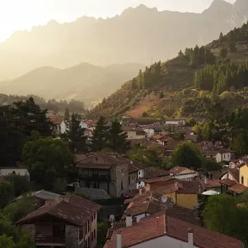 Red-tiled roofs and stone houses of Potes, Cantabria, among pine-covered hills below jagged mountains in hazy golden light.