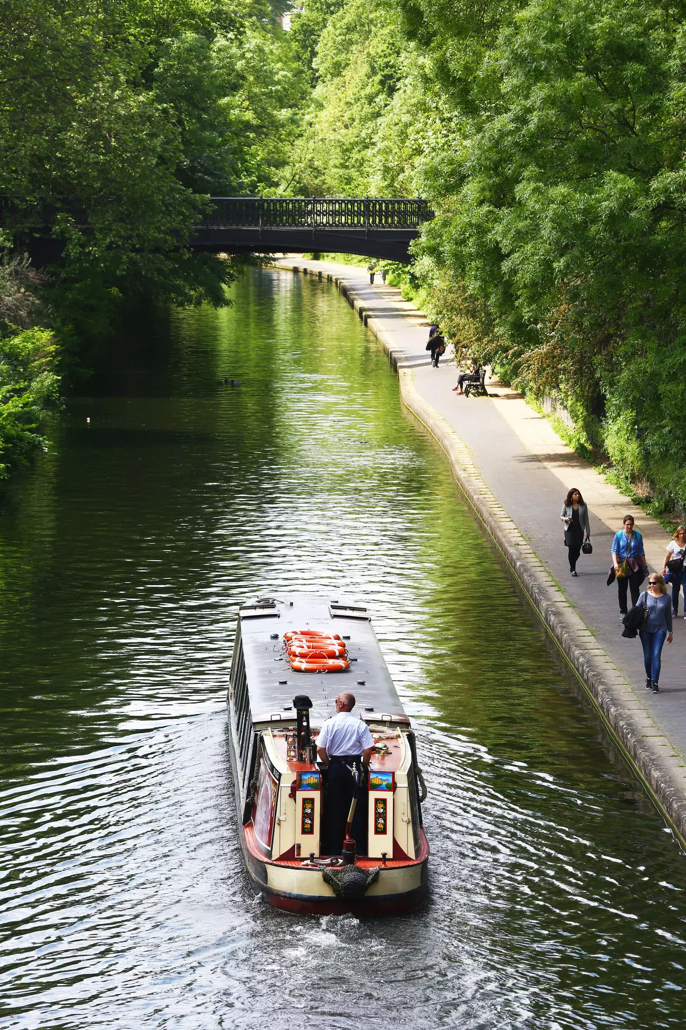 A boat quietly sails on a canal surrounded by trees. Some people walk on the towpath