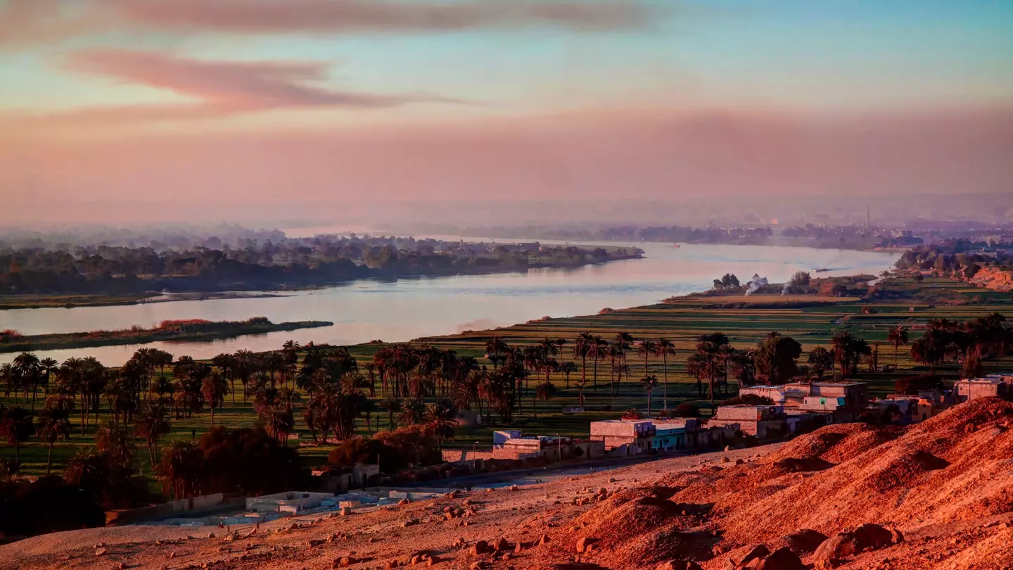Red desolate earth gives way to a small settlement, a flood plain and a wide river as the sun sets.