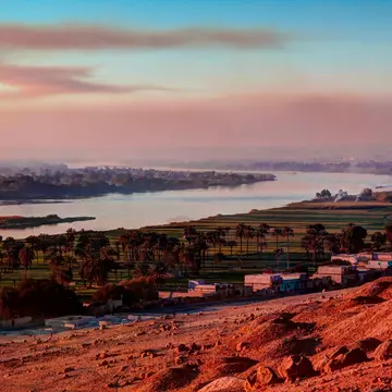Red desolate earth gives way to a small settlement, a flood plain and a wide river as the sun sets.
