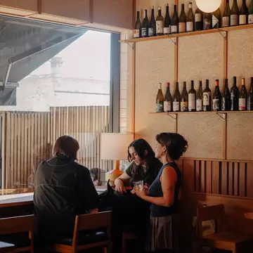 Three people by a large, bright window in a wine bar in Melbourne, Australia. Two are sitting and one is standing with a drink in hand.