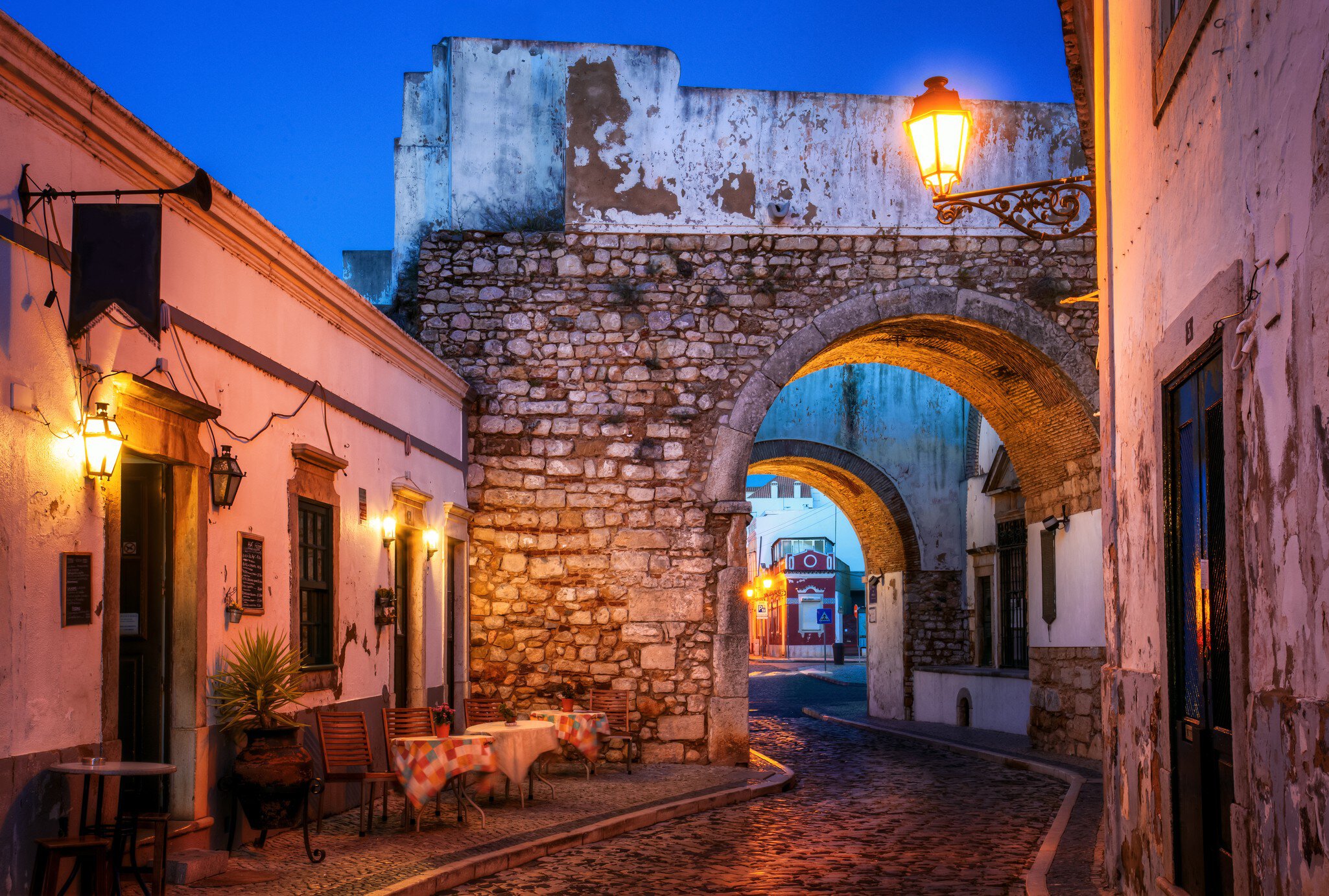 A narrow street in a city in the Algarve in Portugal leads through a stone arch at dusk; there are tables on the sidewalk at the bend by the arch.