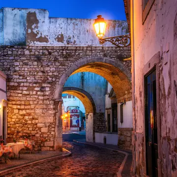 A narrow street in a city in the Algarve in Portugal leads through a stone arch at dusk; there are tables on the sidewalk at the bend by the arch.