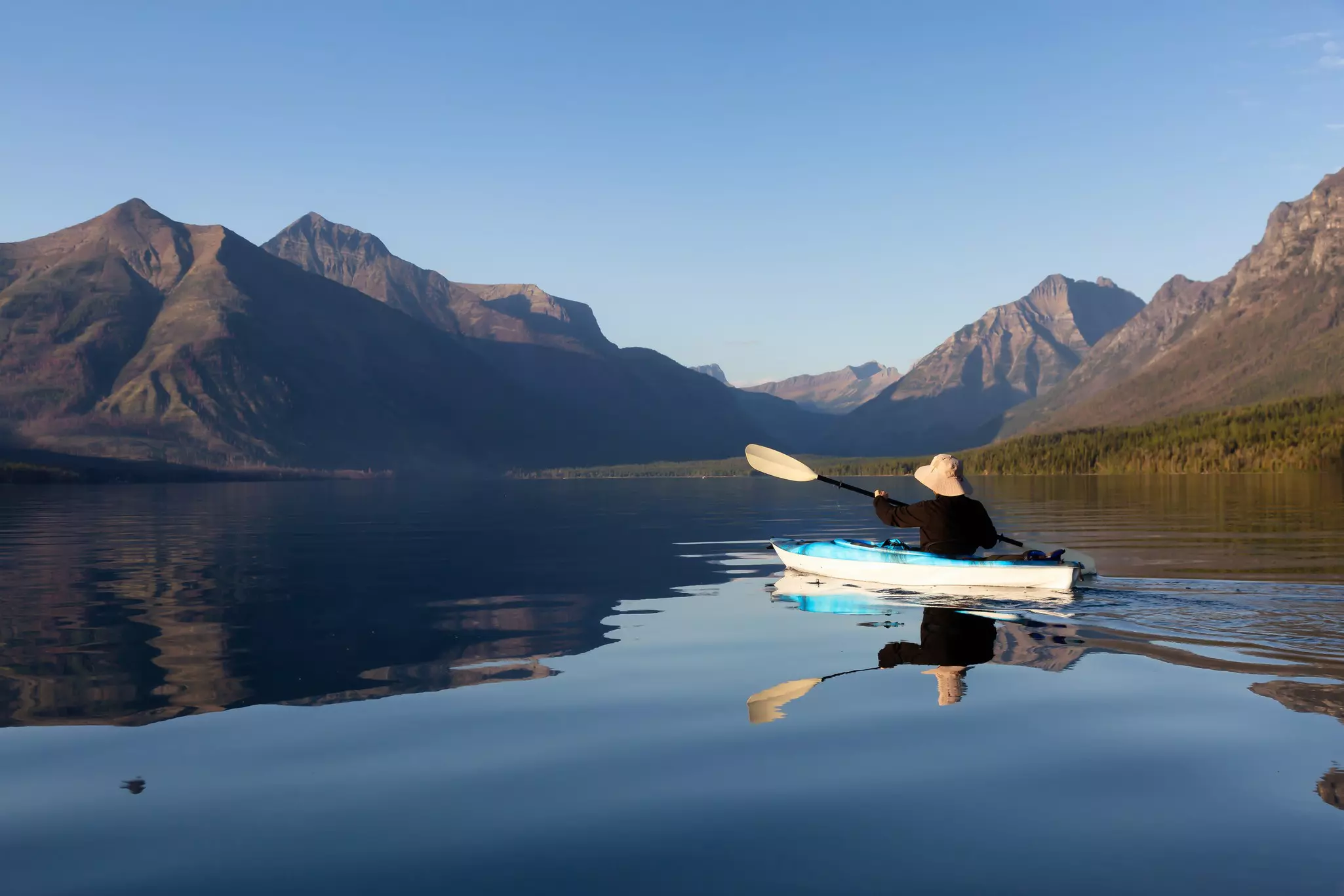 A kayaker paddling on a calm lake with the mountains reflected on the water's surface.