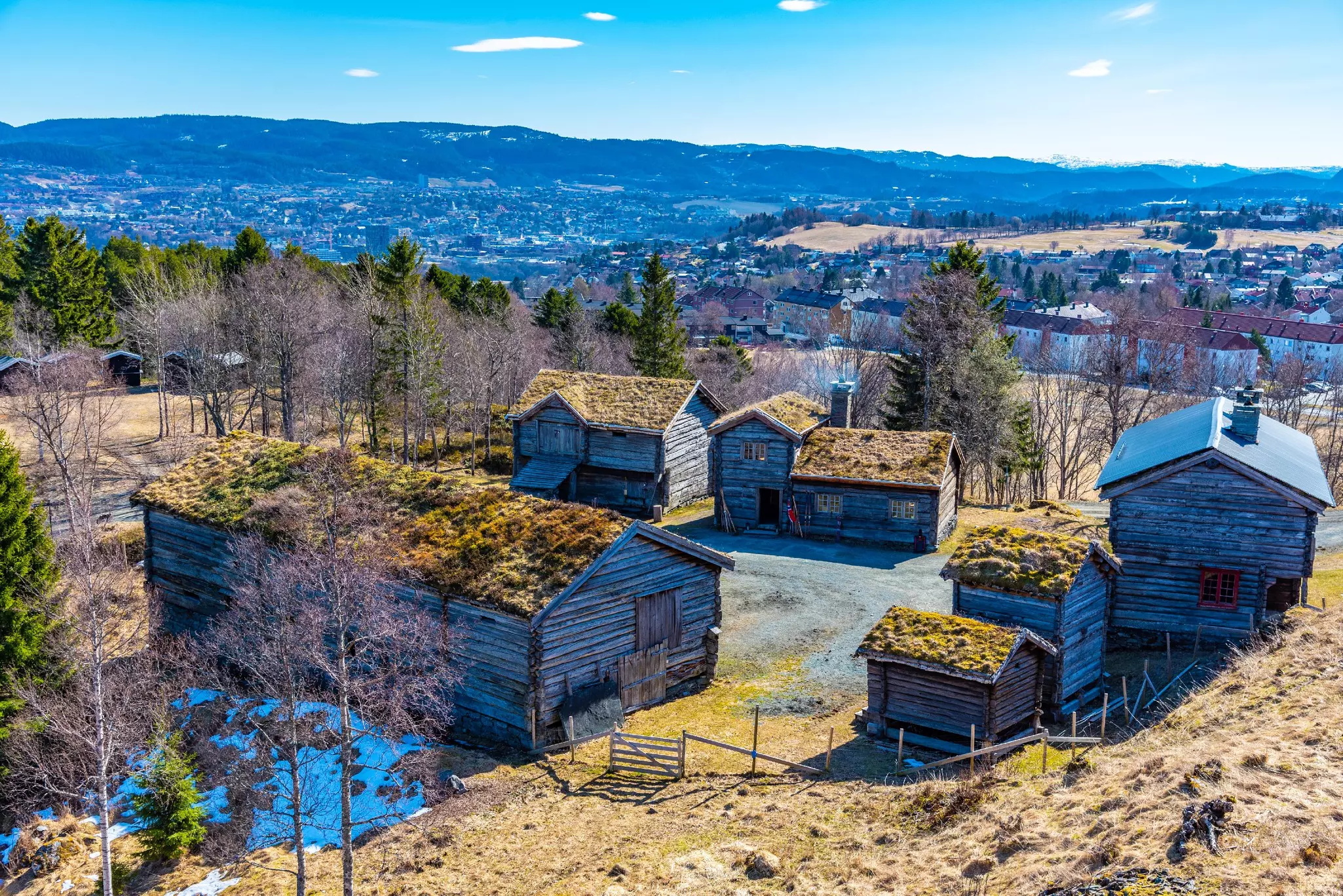 An overhead view of historic wood cabins topped with earth on top of a hill. A city is seen in the valley in the distance.