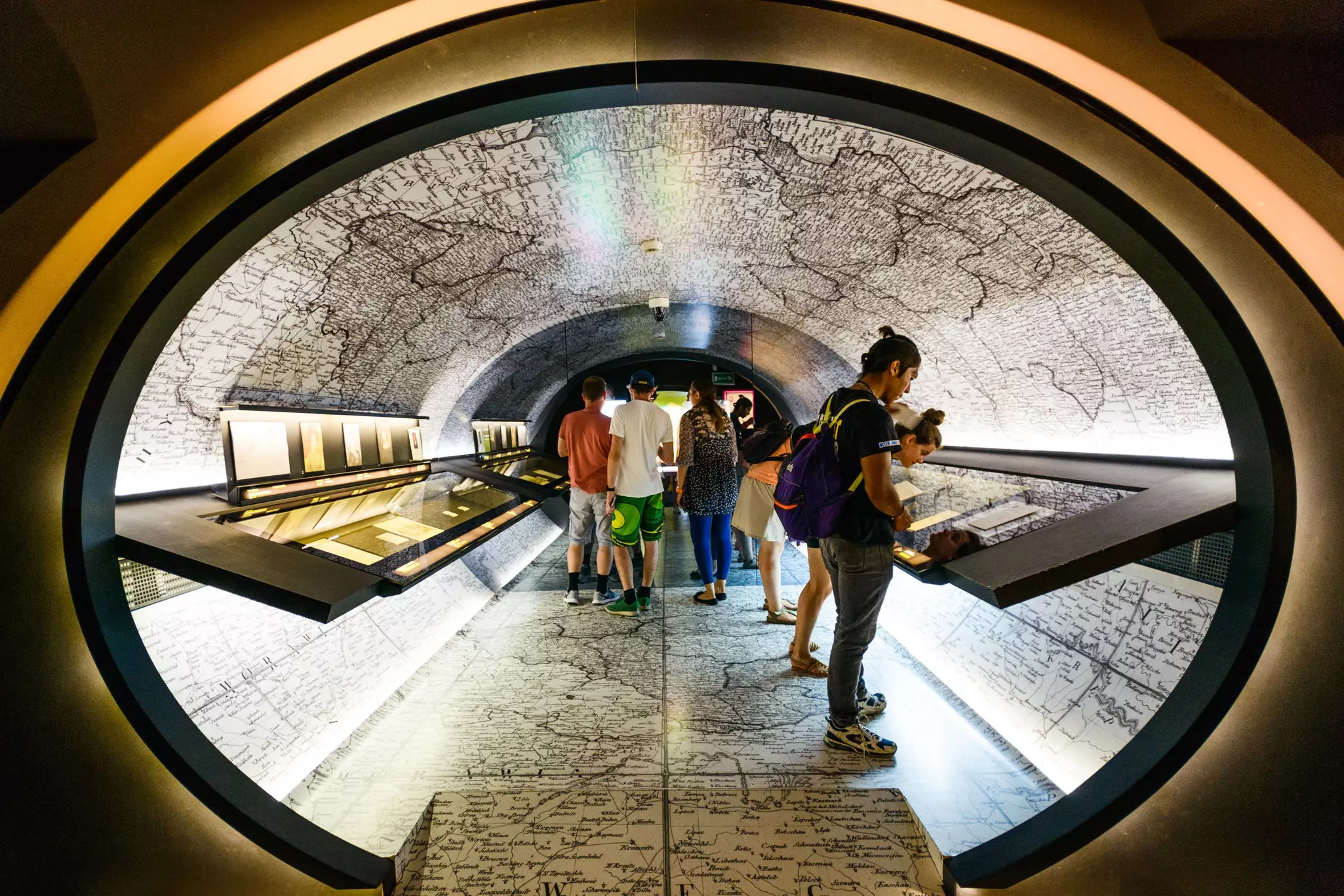People in an oval-shaped hall with glass-encased exhibits and a wall decorated to resemble a map