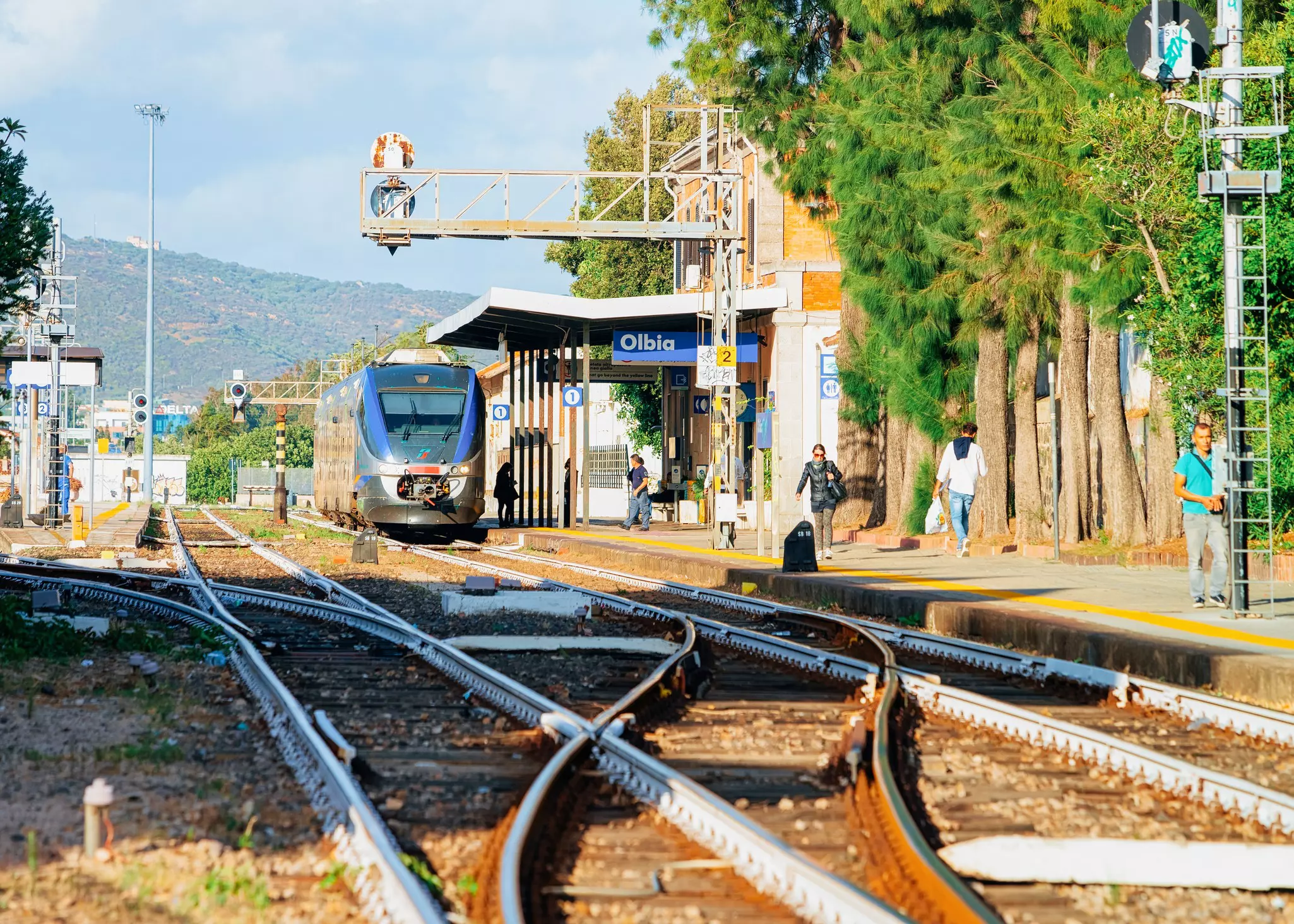 A view of tracks at a train station. People stand on a platform next to the tracks, and a stopped train is visible in the distance.