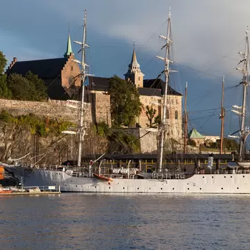 Stone castle with a green spire and square tower on a rocky bluff, a white three-masted ship at the quay.