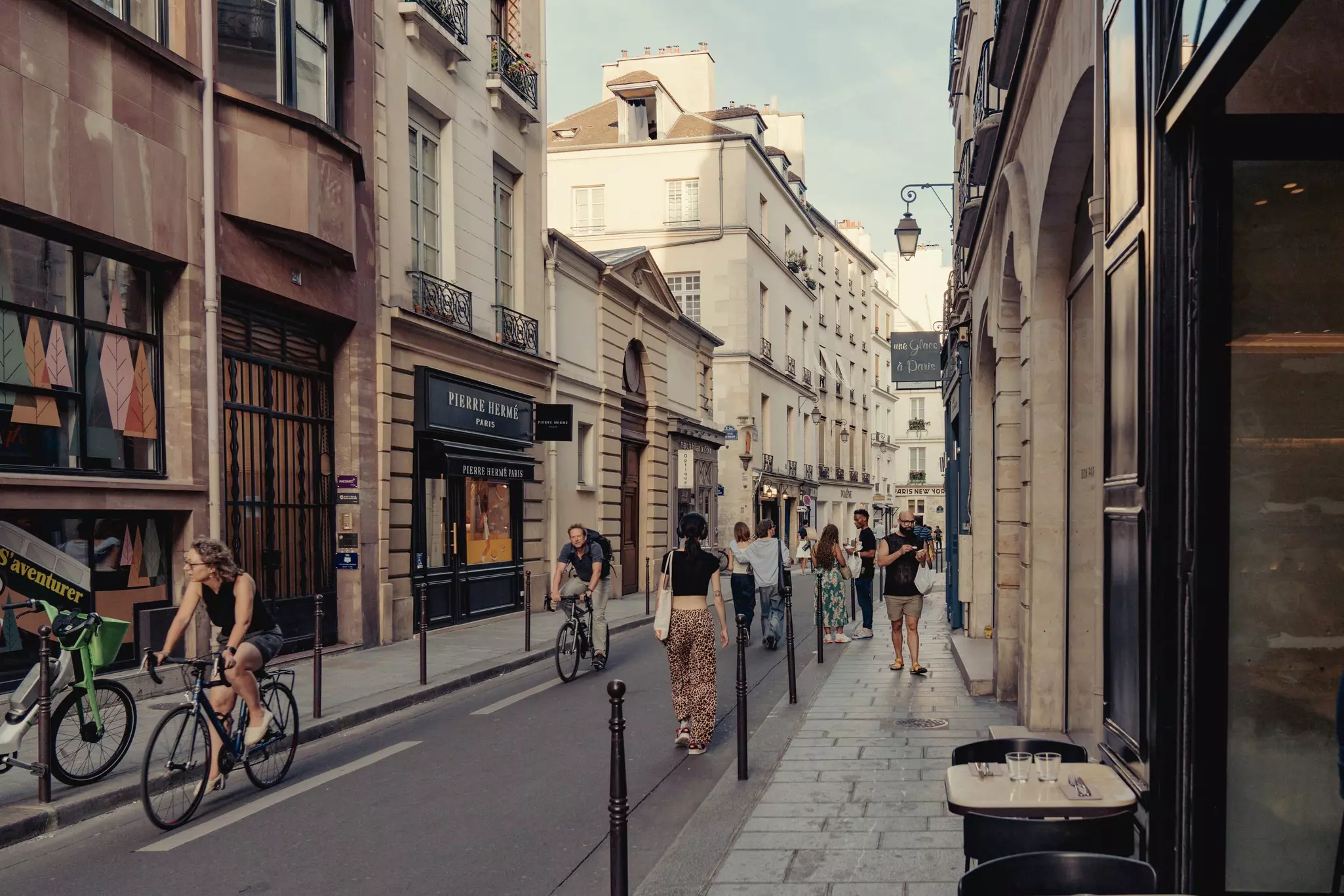 Cyclists and people walking on a narrow street in Paris; there is a cafe table in the foreground and shops on the opposite side of the street.