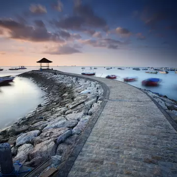Curving stone jetty with a thatched gazebo at its end at dawn, Nusa Dua, Bali, with small boats on still water.