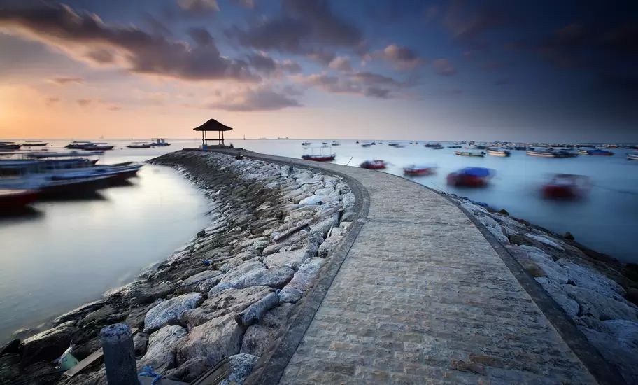 Curving stone jetty with a thatched gazebo at its end at dawn, Nusa Dua, Bali, with small boats on still water.